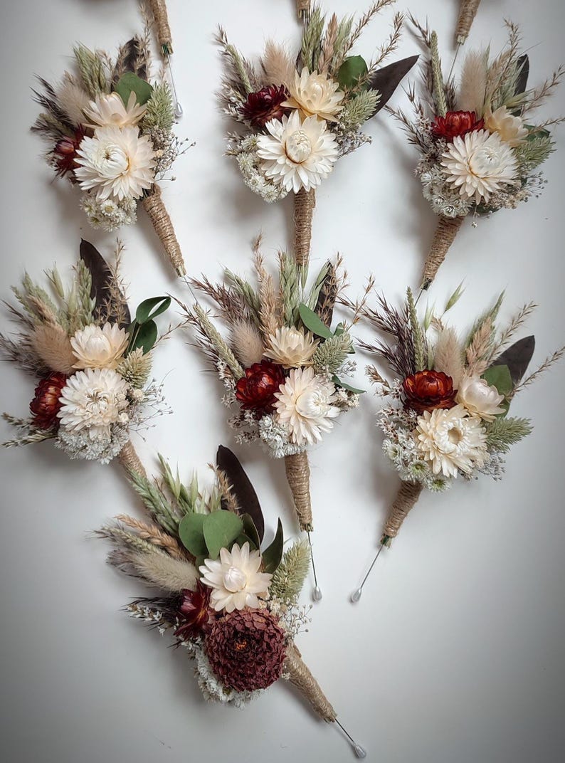 May include: A collection of seven buttonholes featuring dried flowers, including cream and burgundy strawflowers, fluffy pampas grass, green eucalyptus leaves, and wheat stalks. Each arrangement is wrapped in natural jute twine with a metal pin.