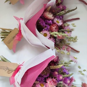 May include: Three bouquets of dried flowers, wrapped in pink and white paper. The bouquets feature a variety of pink, purple, and yellow flowers, with green stems and wheat. Each bouquet has a tag with text.