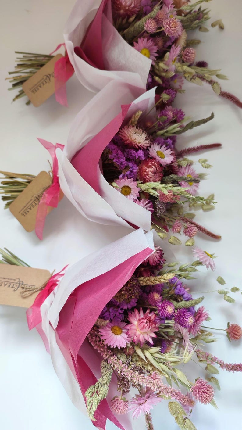 May include: Three bouquets of dried flowers, wrapped in pink and white paper. The bouquets feature a variety of pink, purple, and yellow flowers, with green stems and wheat. Each bouquet has a tag with text.
