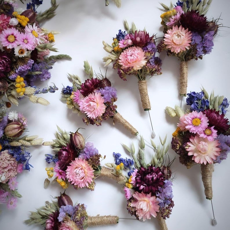 May include: A collection of dried flower buttonholes featuring pink, purple, yellow, and deep burgundy blossoms mixed with dried grasses. Each arrangement is bound with natural twine and includes a metal pin for attachment to clothing.