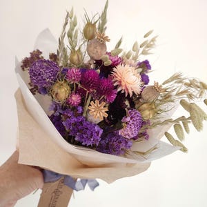 May include: A bouquet of dried flowers in shades of purple, pink, and beige, wrapped in tan paper. The arrangement includes various textures and shapes, with a tag tied with a blue ribbon.