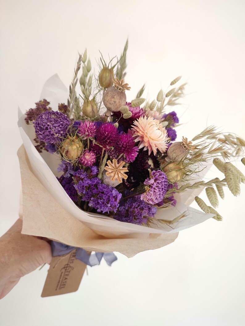 May include: A bouquet of dried flowers in shades of purple, pink, and beige, wrapped in tan paper. The arrangement includes various textures and shapes, with a tag tied with a blue ribbon.