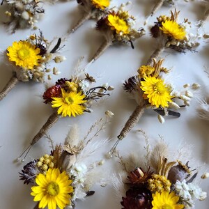 May include: A collection of buttonholes featuring bright yellow strawflowers, dark burgundy dried flowers, white filler flowers, and fluffy pampas grass stems. Each floral arrangement is bound with natural jute twine at the base of the stems.