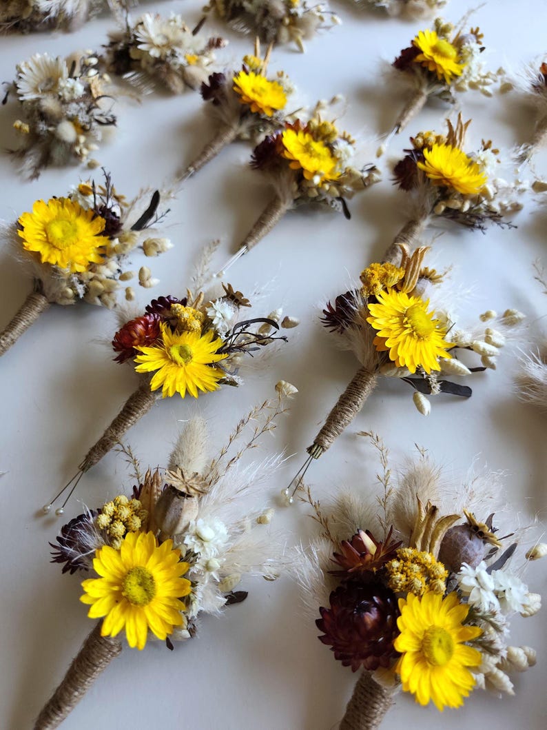 May include: A collection of buttonholes featuring bright yellow strawflowers, dark burgundy dried flowers, white filler flowers, and fluffy pampas grass stems. Each floral arrangement is bound with natural jute twine at the base of the stems.