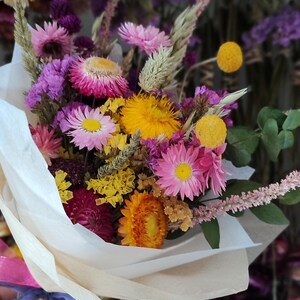 May include: A colorful bouquet of dried flowers, including pink, yellow, and purple blooms, wrapped in white paper. The arrangement features various textures and shapes, creating a vibrant and natural aesthetic.