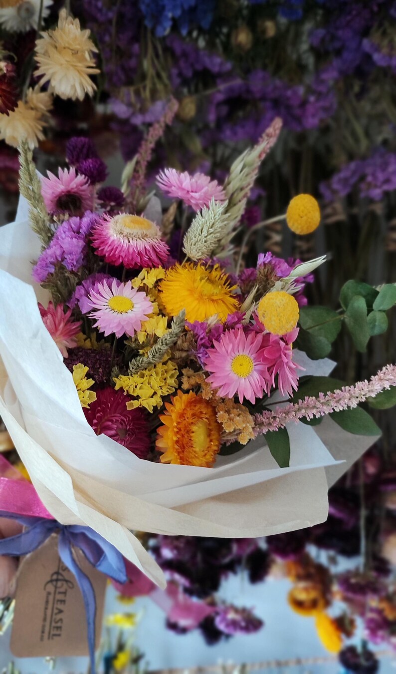 May include: A colorful bouquet of dried flowers, including pink, yellow, and purple blooms, wrapped in white paper. The arrangement features various textures and shapes, creating a vibrant and natural aesthetic.