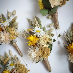 May include: A collection of four buttonholes featuring dried flowers in shades of yellow, cream, and green. Each arrangement includes daisies, strawflowers, and wheat stalks, with stems wrapped in natural twine and finished with a silver pin.