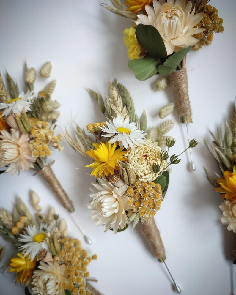 May include: A collection of four buttonholes featuring dried flowers in shades of yellow, cream, and green. Each arrangement includes daisies, strawflowers, and wheat stalks, with stems wrapped in natural twine and finished with a silver pin.