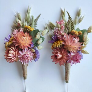 May include: Two floral buttonholes featuring dried flowers in shades of peach, pink, yellow, and purple, accented with green foliage and wheat stalks. The stems are wrapped in natural jute twine and each includes a long silver metal pin at the base.