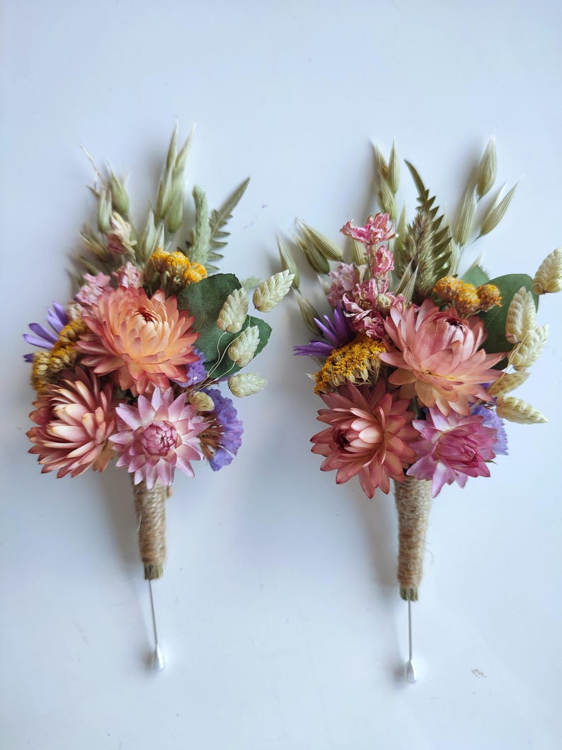 May include: Two floral buttonholes featuring dried flowers in shades of peach, pink, yellow, and purple, accented with green foliage and wheat stalks. The stems are wrapped in natural jute twine and each includes a long silver metal pin at the base.