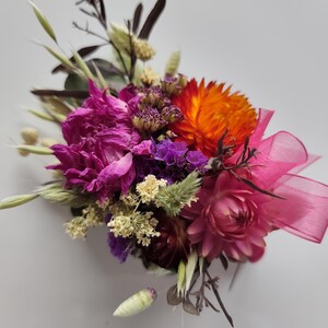 May include: A floral buttonhole featuring dried flowers in vibrant shades of magenta, bright orange, and deep purple, accented with small cream-coloured sprigs, green grasses, and a translucent pink ribbon tied in a bow on the right side.