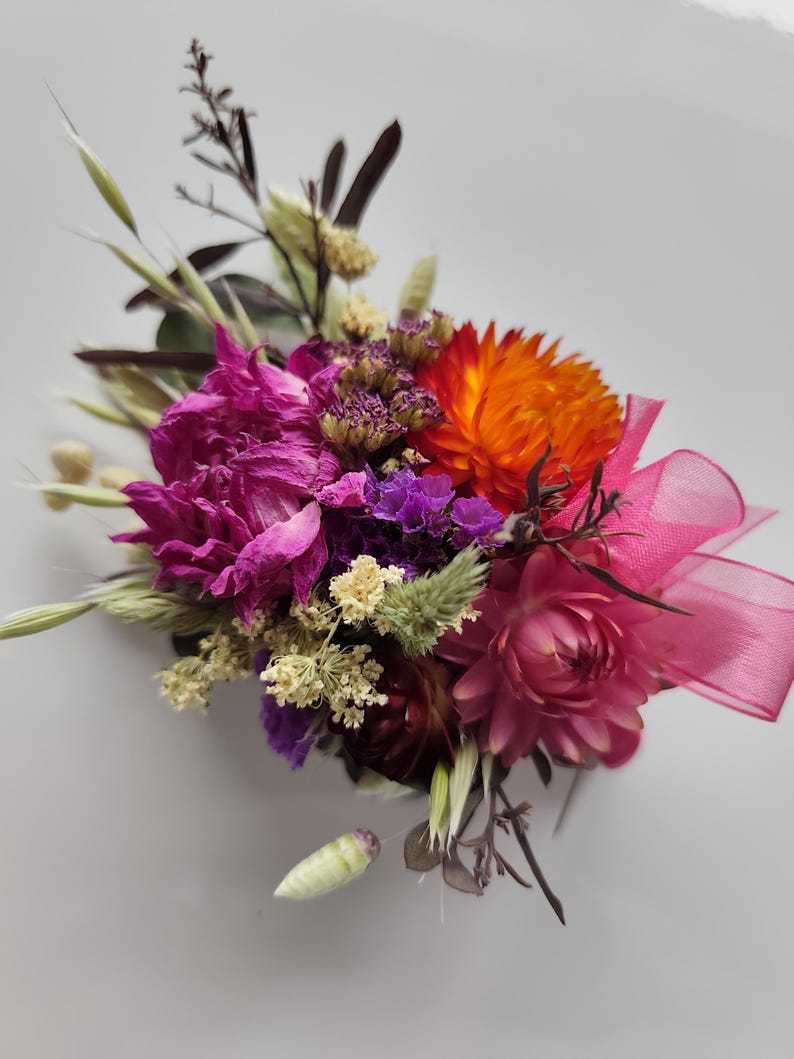 May include: A floral buttonhole featuring dried flowers in vibrant shades of magenta, bright orange, and deep purple, accented with small cream-coloured sprigs, green grasses, and a translucent pink ribbon tied in a bow on the right side.