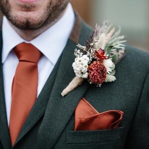 May include: A dark green suit jacket with a white collared shirt, a solid burnt orange necktie, and a matching burnt orange pocket square. A boutonniere made of dried white, red, and brown flowers is pinned to the lapel with jute twine.
