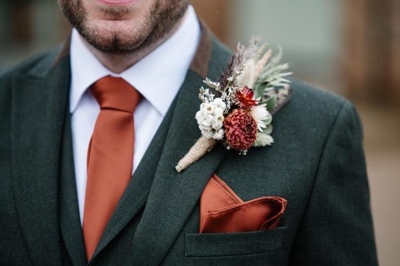 May include: A dark green suit jacket with a white collared shirt, a solid burnt orange necktie, and a matching burnt orange pocket square. A boutonniere made of dried white, red, and brown flowers is pinned to the lapel with jute twine.