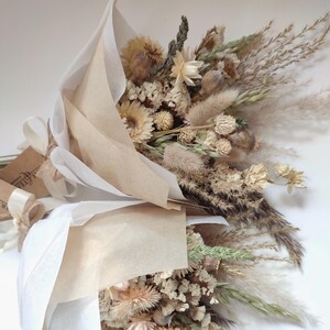 May include: Two bouquets of dried flowers, wrapped in beige paper and tied with cream-colored ribbons. The bouquets feature a variety of beige, tan, and green dried flowers and grasses. The arrangement is a natural, rustic style.