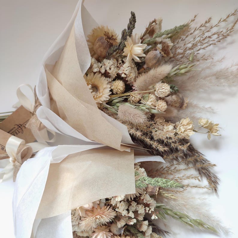 May include: Two bouquets of dried flowers, wrapped in beige paper and tied with cream-colored ribbons. The bouquets feature a variety of beige, tan, and green dried flowers and grasses. The arrangement is a natural, rustic style.