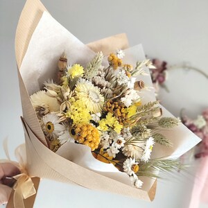 May include: A bouquet of dried flowers in shades of yellow, white, and beige, wrapped in brown paper and tied with a beige ribbon. The bouquet includes daisies, wheat, and other dried floral elements. The background is a soft, neutral color.