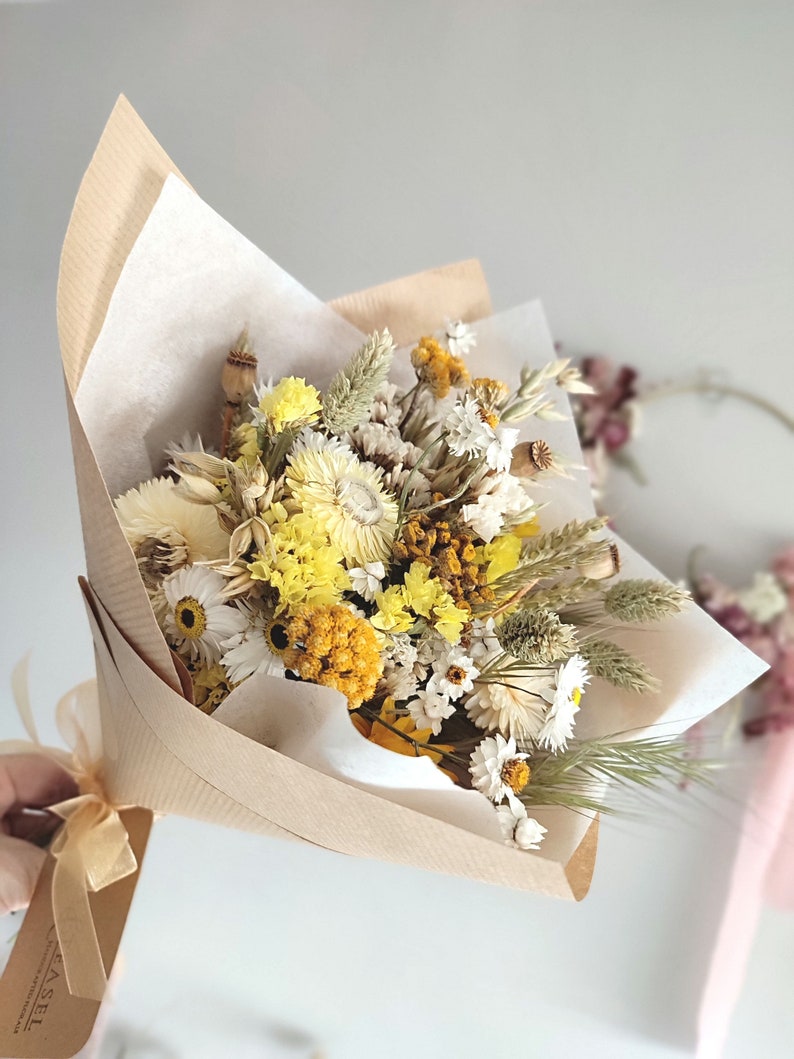 May include: A bouquet of dried flowers in shades of yellow, white, and beige, wrapped in brown paper and tied with a beige ribbon. The bouquet includes daisies, wheat, and other dried floral elements. The background is a soft, neutral color.
