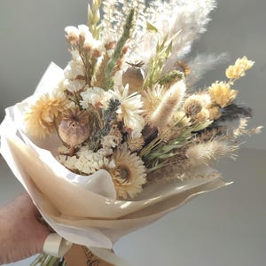 May include: A bouquet of dried flowers in shades of cream, beige, and yellow, wrapped in white and tan paper. The bouquet includes various textures of dried grasses, seed pods, and delicate blooms, tied with a white ribbon and a brown tag.