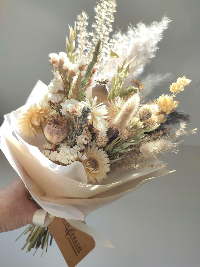May include: A bouquet of dried flowers in shades of cream, beige, and yellow, wrapped in white and tan paper. The bouquet includes various textures of dried grasses, seed pods, and delicate blooms, tied with a white ribbon and a brown tag.