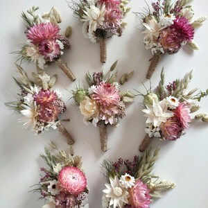 May include: Eight floral buttonholes featuring dried flowers in shades of pink, cream, and green. Each arrangement includes strawflowers, grasses, and small white blossoms, with stems wrapped in natural jute twine for a secure, textured finish.