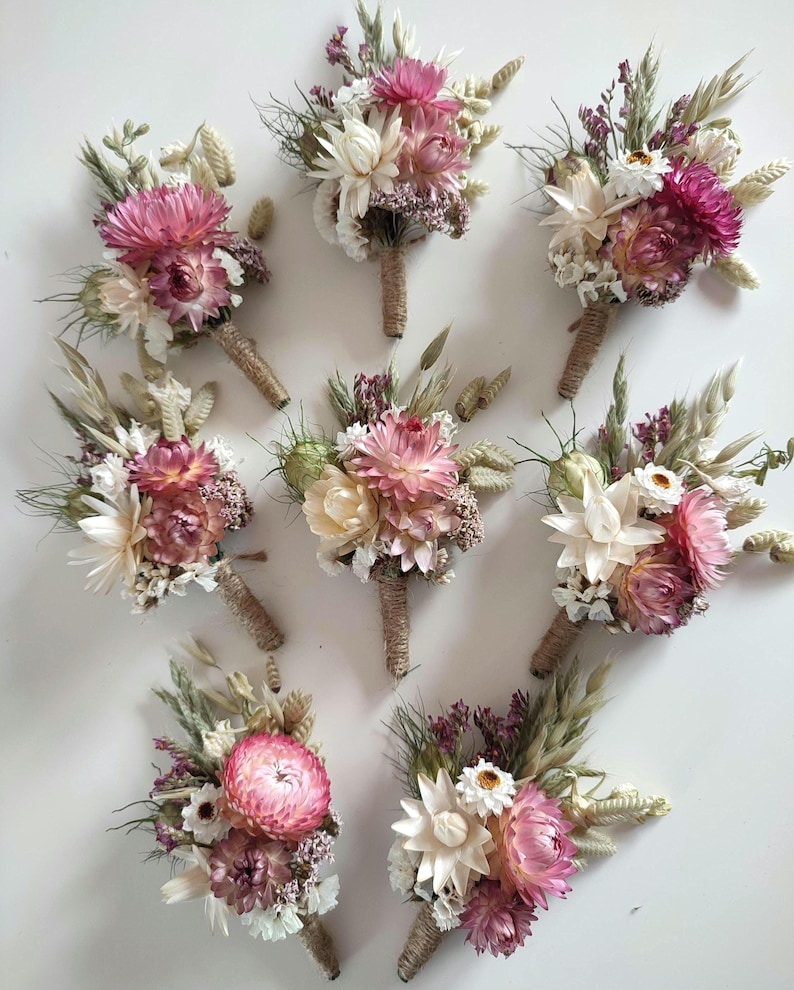 May include: Eight floral buttonholes featuring dried flowers in shades of pink, cream, and green. Each arrangement includes strawflowers, grasses, and small white blossoms, with stems wrapped in natural jute twine for a secure, textured finish.