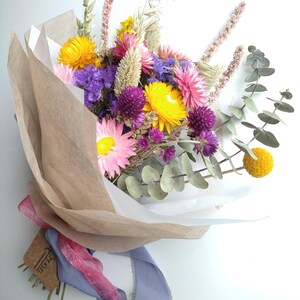 May include: A vibrant bouquet of dried flowers, featuring yellow, pink, and purple blooms, wrapped in brown and white paper. The bouquet is tied with a pink and blue ribbon, and includes sprigs of eucalyptus. A tag with the word "TEASEL" is attached.