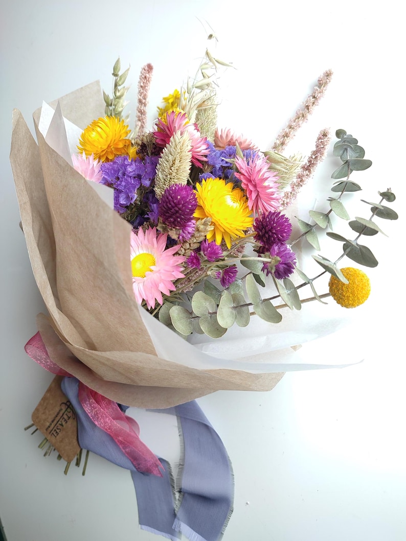 May include: A vibrant bouquet of dried flowers, featuring yellow, pink, and purple blooms, wrapped in brown and white paper. The bouquet is tied with a pink and blue ribbon, and includes sprigs of eucalyptus. A tag with the word "TEASEL" is attached.