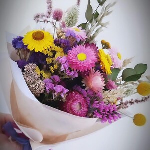 May include: A colorful bouquet of dried flowers, including yellow, pink, purple, and white blooms. The flowers are wrapped in tan paper and tied with a blue and purple ribbon. The bouquet is held against a white background.