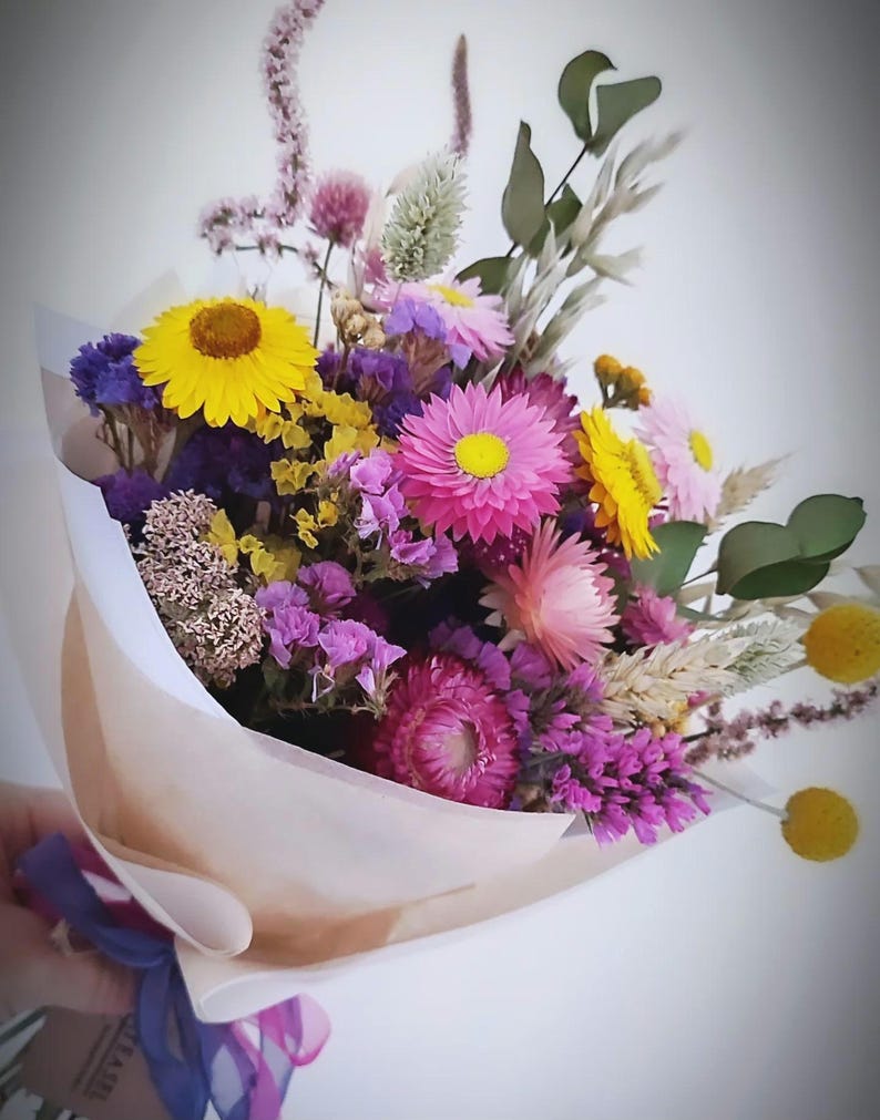 May include: A colorful bouquet of dried flowers, including yellow, pink, purple, and white blooms. The flowers are wrapped in tan paper and tied with a blue and purple ribbon. The bouquet is held against a white background.