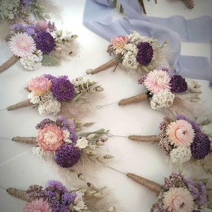 May include: A collection of eight buttonholes featuring dried flowers in shades of purple, pink, and cream. Each arrangement includes strawflowers, pampas grass, and other dried botanicals, with stems wrapped in twine and a long silver pin attached.