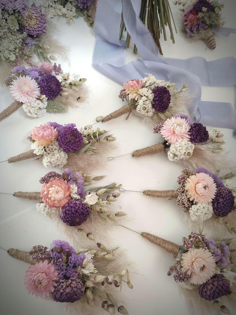 May include: A collection of eight buttonholes featuring dried flowers in shades of purple, pink, and cream. Each arrangement includes strawflowers, pampas grass, and other dried botanicals, with stems wrapped in twine and a long silver pin attached.