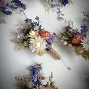 May include: A collection of buttonholes made from dried flowers including white, peach, and pink strawflowers, blue delphiniums, and wheat stalks. Each arrangement is bound with natural jute twine and features a long silver pin for attachment.
