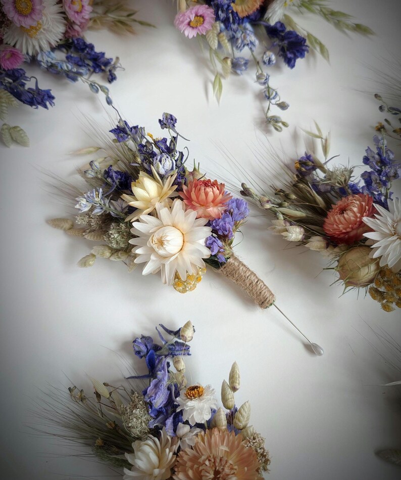 May include: A collection of buttonholes made from dried flowers including white, peach, and pink strawflowers, blue delphiniums, and wheat stalks. Each arrangement is bound with natural jute twine and features a long silver pin for attachment.