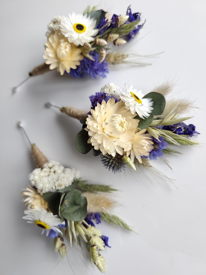 May include: Three floral buttonholes featuring cream-coloured strawflowers, white daisies with dark centres, blue statice, green eucalyptus leaves, dried wheat stalks, and fluffy beige pampas grass, all wrapped in natural jute twine with a silver pin.