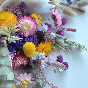 May include: A vibrant bouquet of dried flowers, featuring a mix of textures and colors. The arrangement includes yellow, pink, purple, and blue blooms, with accents of green foliage. The bouquet is wrapped in a neutral-toned paper.