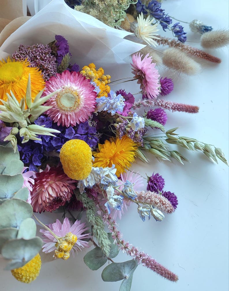 May include: A vibrant bouquet of dried flowers, featuring a mix of textures and colors. The arrangement includes yellow, pink, purple, and blue blooms, with accents of green foliage. The bouquet is wrapped in a neutral-toned paper.