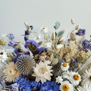 May include: A bouquet of dried flowers in shades of blue and white. The bouquet includes white daisies, blue delphiniums, and other dried flowers.