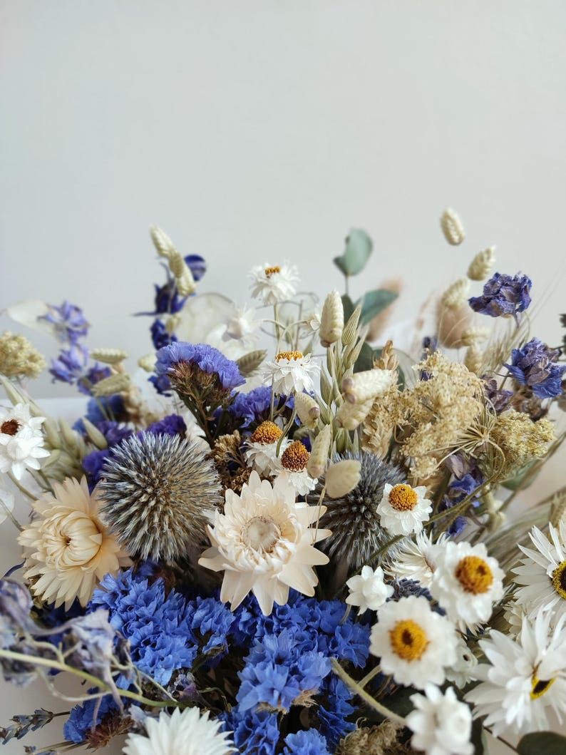 May include: A bouquet of dried flowers in shades of blue and white. The bouquet includes white daisies, blue delphiniums, and other dried flowers.
