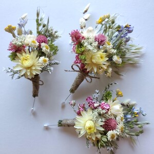May include: Three boutonnieres featuring dried flowers in shades of yellow, pink, white, and green. Each arrangement includes a large central yellow strawflower, small pink clover-like blooms, and wispy grasses, all wrapped in brown twine with a pin.