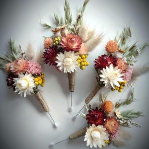 May include: Four floral buttonholes featuring dried flowers in shades of burgundy, cream, peach, and yellow, accented with green grasses and fluffy beige plumes. Each arrangement is wrapped in natural jute twine and includes a long metal pin.