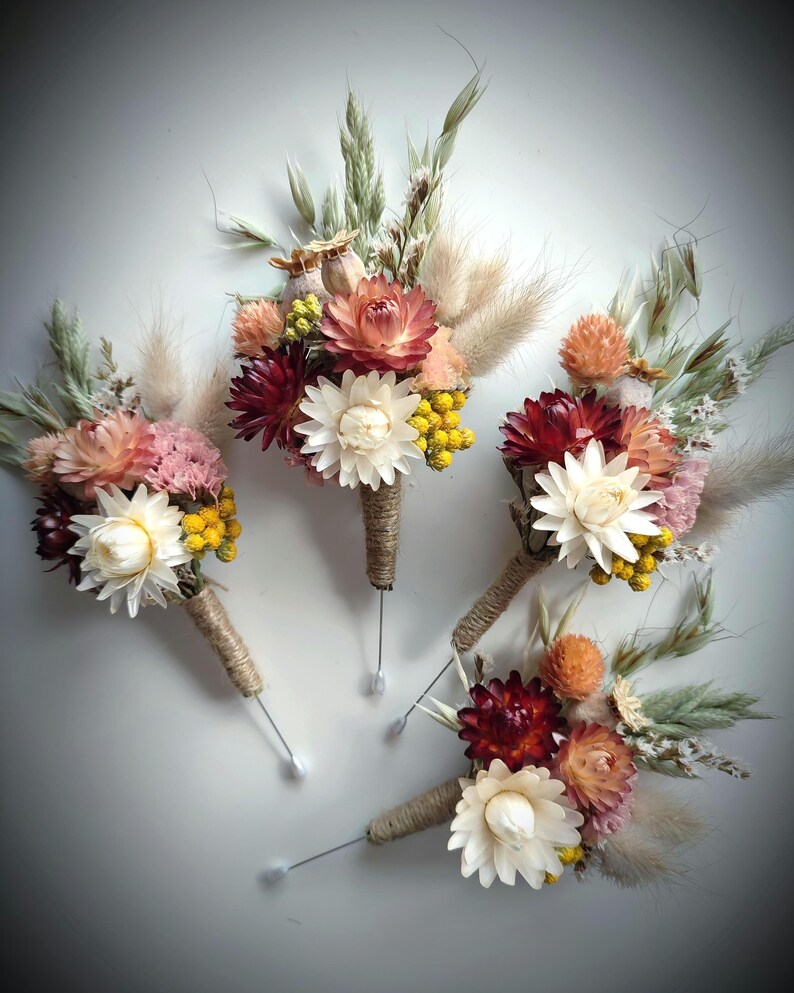 May include: Four floral buttonholes featuring dried flowers in shades of burgundy, cream, peach, and yellow, accented with green grasses and fluffy beige plumes. Each arrangement is wrapped in natural jute twine and includes a long metal pin.
