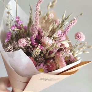 May include: A bouquet of dried flowers in shades of pink, purple, and beige, wrapped in brown paper and tied with a pink ribbon. The arrangement includes various flower types and grasses.
