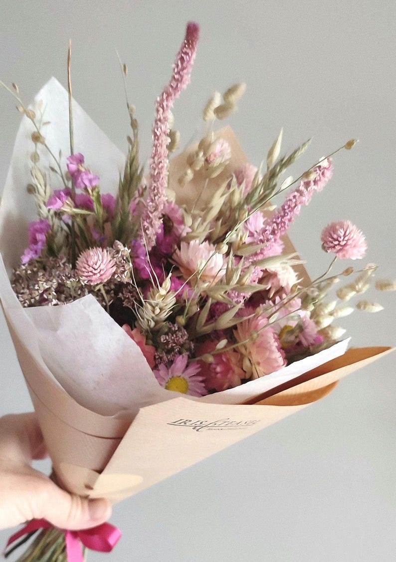 May include: A bouquet of dried flowers in shades of pink, purple, and beige, wrapped in brown paper and tied with a pink ribbon. The arrangement includes various flower types and grasses.