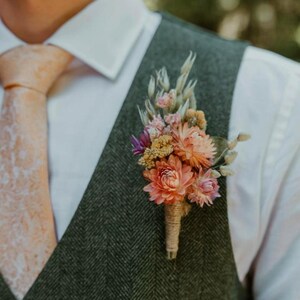 May include: A buttonhole featuring dried flowers in shades of peach, pink, and cream, wrapped in twine. The floral arrangement is pinned to a dark green herringbone waistcoat worn over a white dress shirt with a light peach patterned necktie.