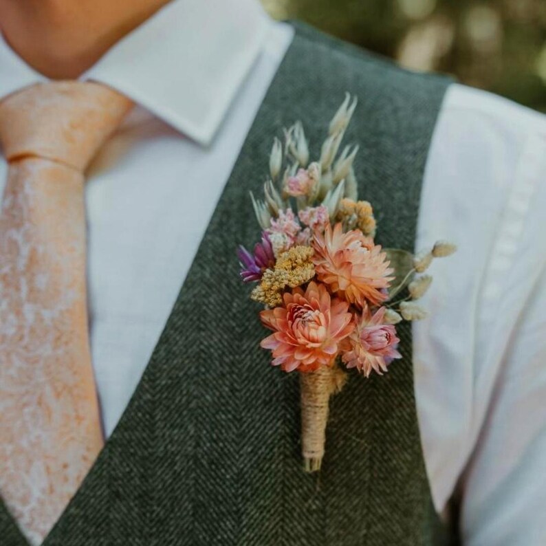 May include: A buttonhole featuring dried flowers in shades of peach, pink, and cream, wrapped in twine. The floral arrangement is pinned to a dark green herringbone waistcoat worn over a white dress shirt with a light peach patterned necktie.