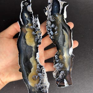 Puede incluir: Dos rodajas de ágata de forma irregular con una mezcla de tonos negros, blancos y amarillos. Los bordes están revestidos con un material cristalino gris-blanco, creando un contraste llamativo. Las piezas de piedra natural se muestran sobre un fondo oscuro.