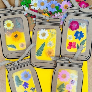 May include: Six brown paper mason jar ornaments with pressed flowers inside. The jars are decorated with black outlines and have a string loop at the top. The jars are filled with a variety of dried flowers, including daisies, lavender, and pink flowers. The jars are displayed on a yellow background.