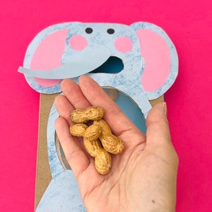 Könnte beinhalten: Eine Hand hält eine Handvoll Erdnüsse vor einem blauen und rosa Papier-Elefanten-Ausschnitt. Der Elefant hat ein Loch in der Mitte und eine Klappe für seinen Rüssel.