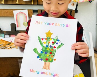 May include: A child holds a handmade Christmas card with a green handprint tree decorated with colorful lights and a gold star. The card reads "May your days be Merry and bright".